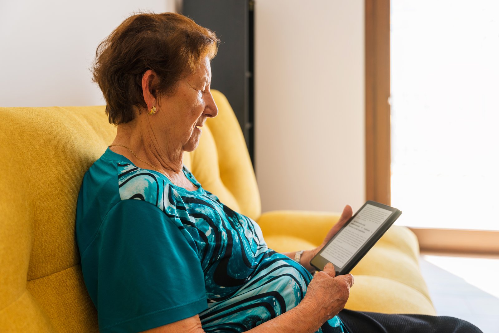 An older woman resting on a yellow sofa at home, reading on a tablet