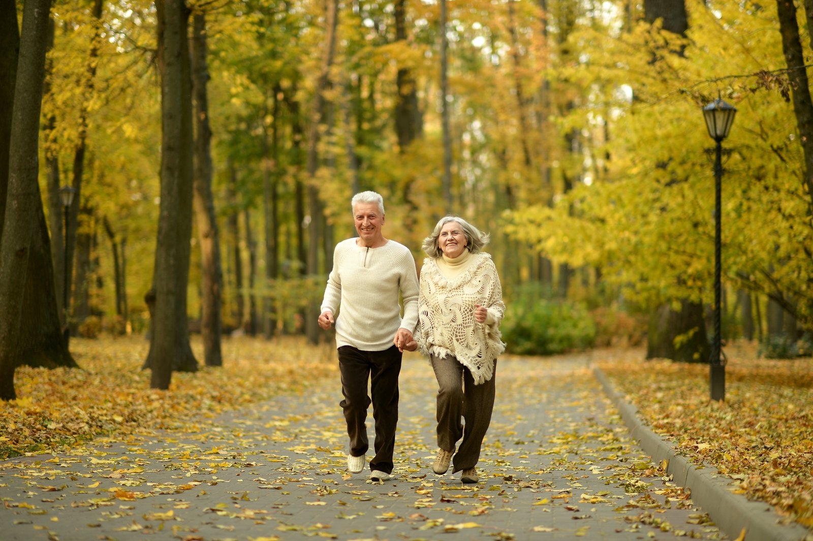An older couple holding hands on a brisk walk along a leaf-covered park path in autumn