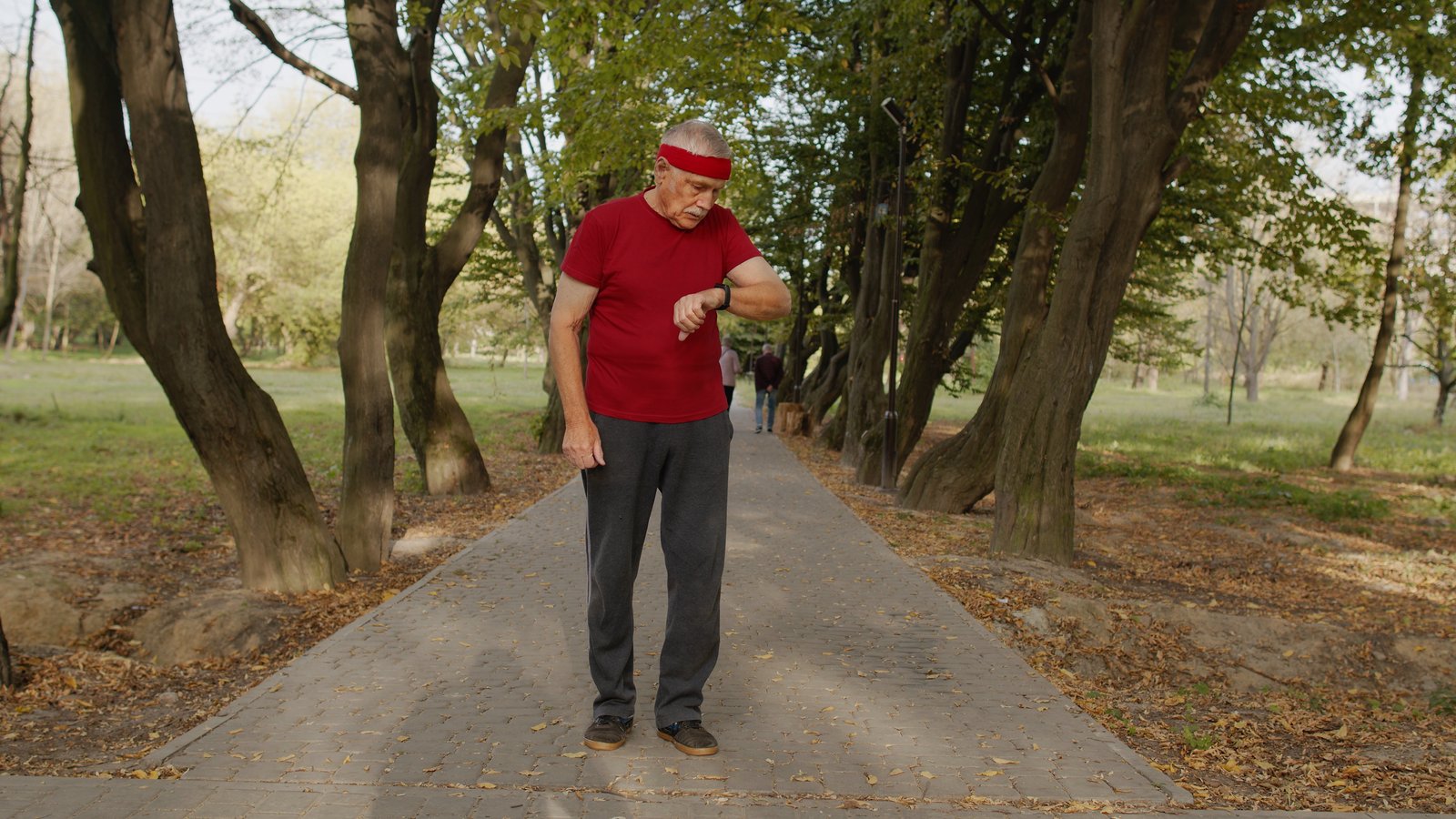 An older man in a red shirt out for a walk on a tree-lined park path