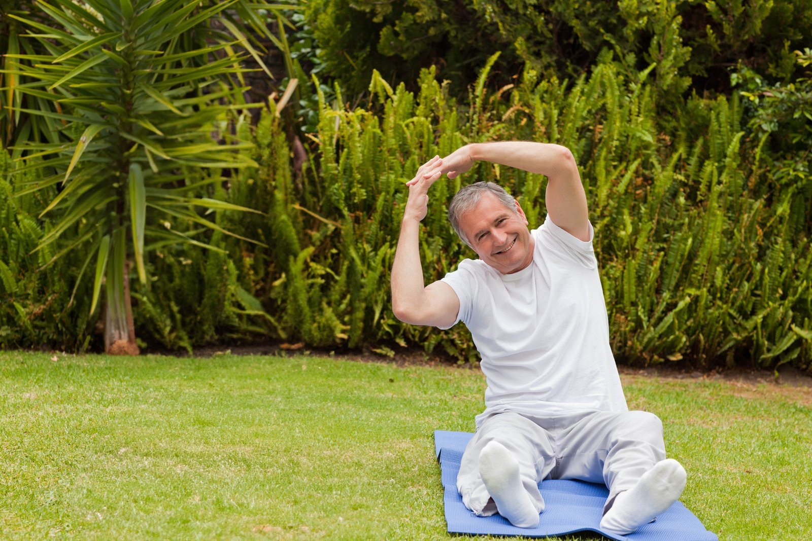 An older man doing a gentle side stretch on a mat in a garden